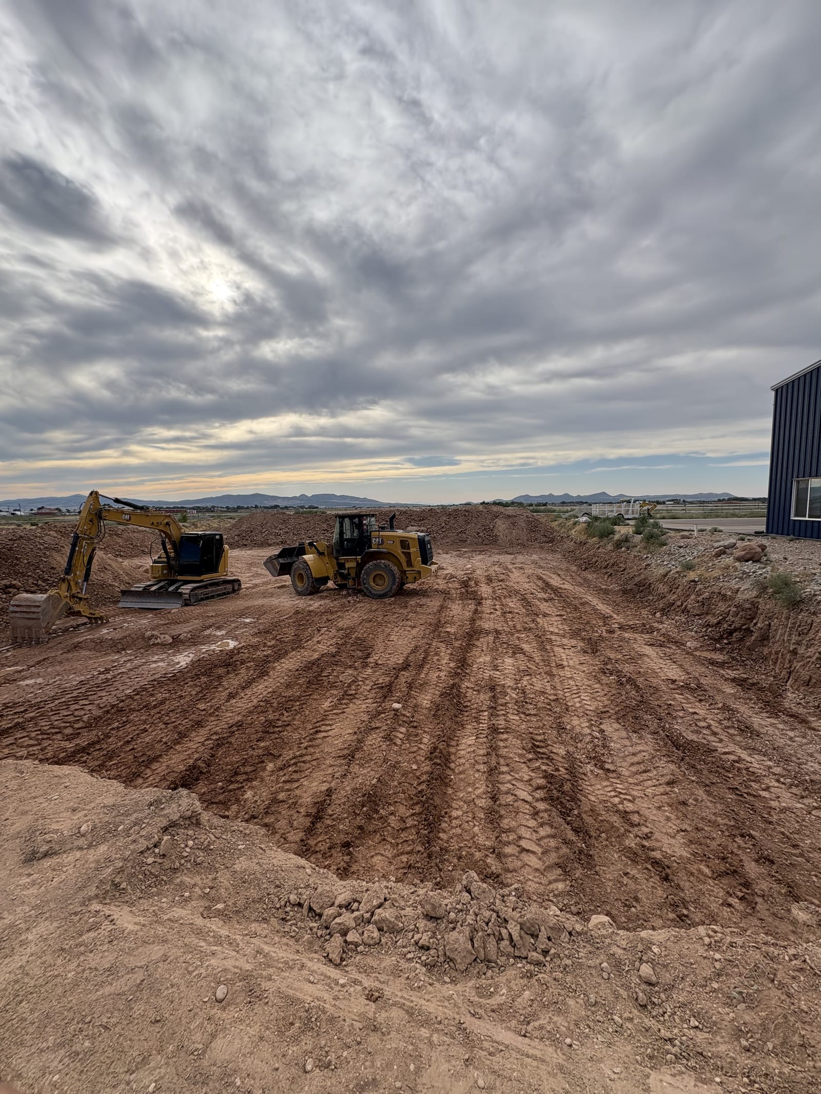 Roxor South CAT excavator and wheel loader staged on a Southern Utah over-excavation pad, with deep roller compaction tracks running across freshly worked structural fill under an overcast desert sky