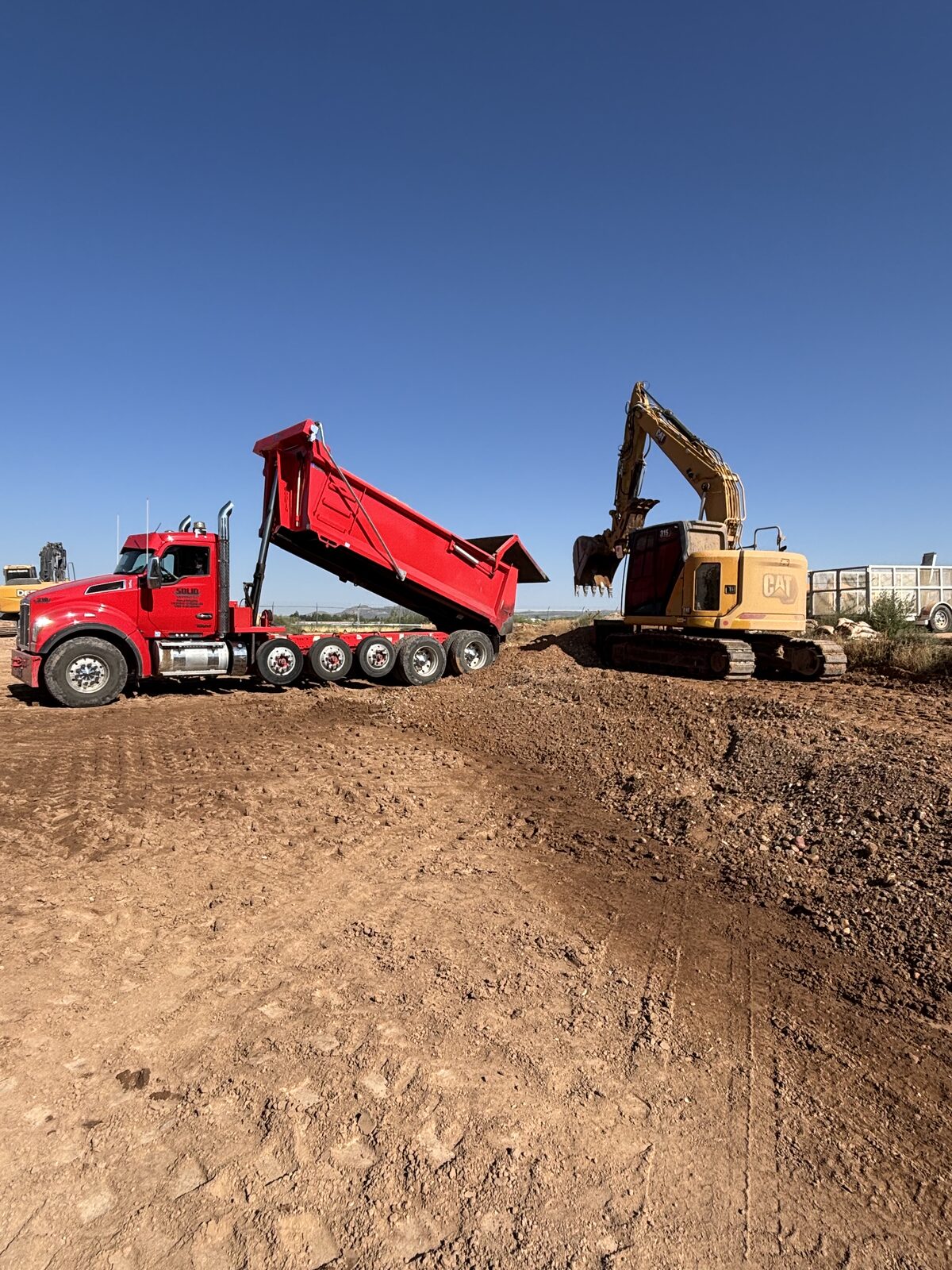 Roxor South red dump truck unloading fill alongside a CAT excavator on a Southern Utah job site