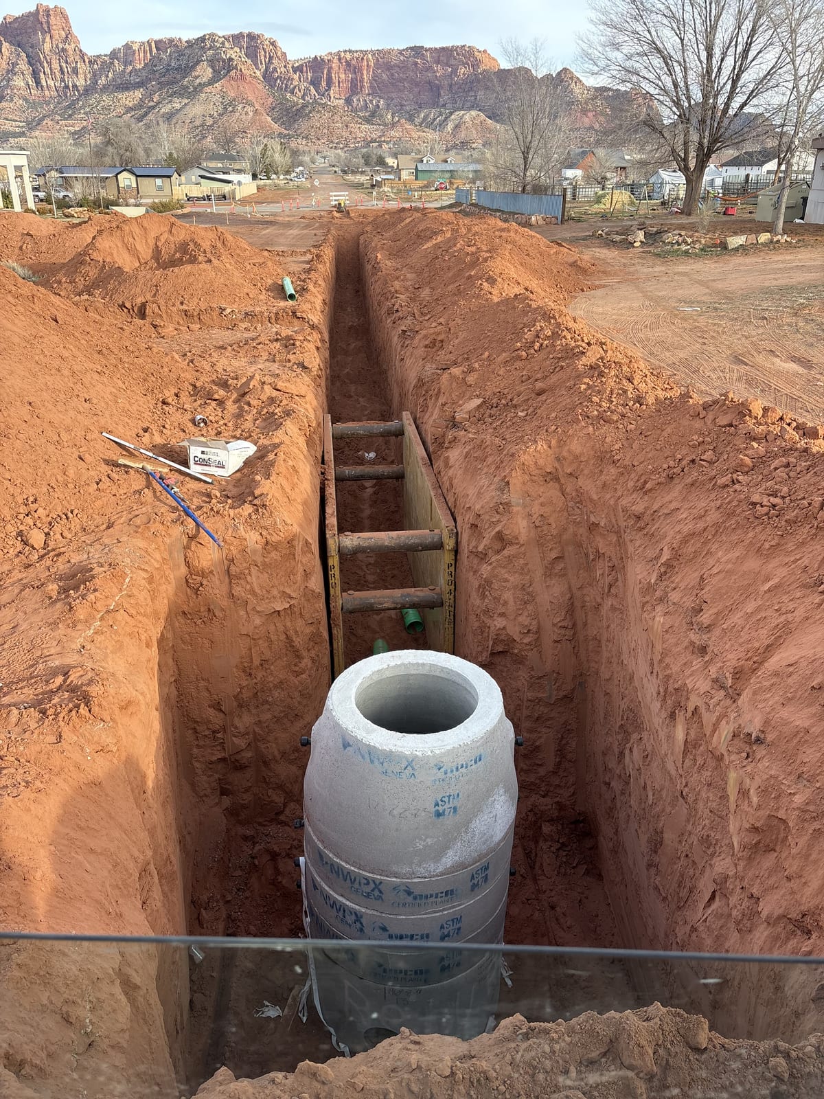 Large precast concrete utility pipe / manhole barrel section staged on wood cribbing inside a deep, square-cut red-dirt trench, Southern Utah rooftops and red rock cliffs in the background — Roxor South underground utilities job in Apple Valley